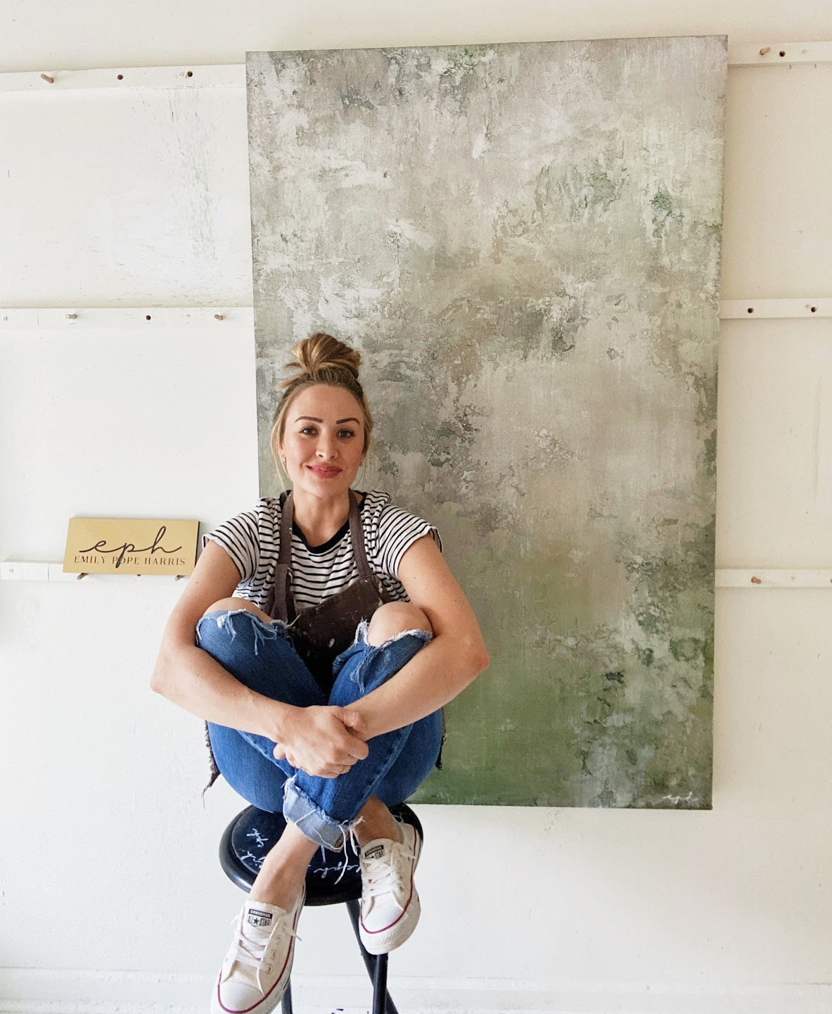 Emily is sitting on a stool in her studio infront of a green and white with grays tone commissioned piece that is on the large scale to a fine art piece. There is texture, depth, and layers of plaster, that make this piece special. 