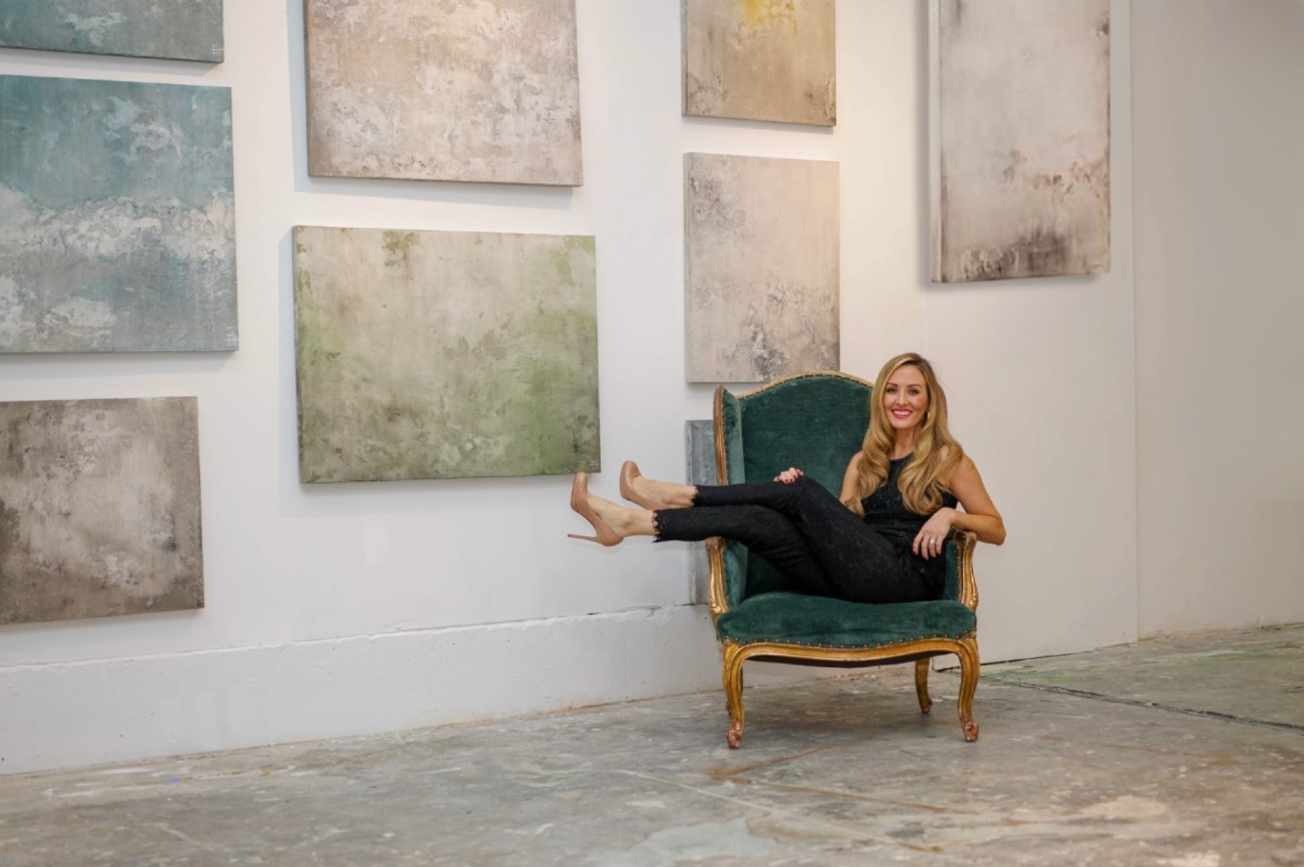 Emily Pope Harris seated in an art gallery with her textured plaster fine art displayed on birch wood panels behind her.