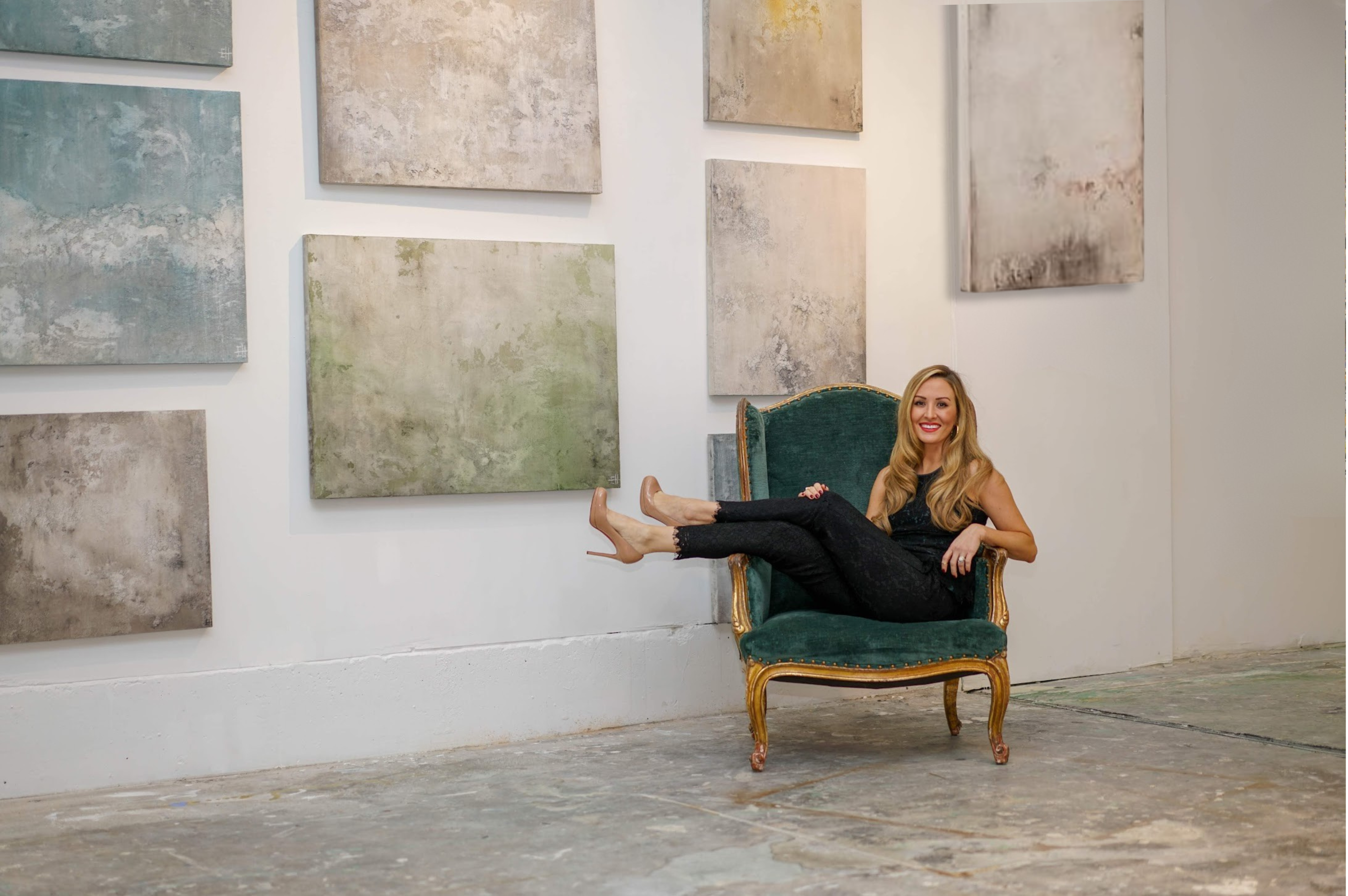 Emily seated at an art gallery with eight heavily textured paintings on birch wood panels displayed behind her in various colorways.