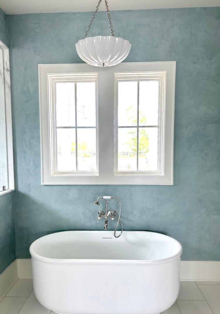 Light blue Venetian plaster wall in a Ravenel, SC master bathroom with burnish and polish finish, looking out to the side yard with horses, and a bathtub in front, created by Emily Pope Harris with Jackson Built Custom Homes.
